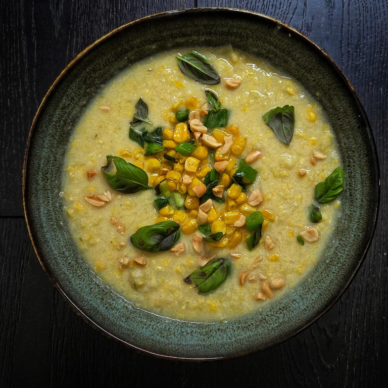 eine cremige gelbliche suppe in einer dunklen schale, mit erkennbaren Maisstückchen und einem Mais-Chili-Topping sowie einzelnen Basilikumblättchen